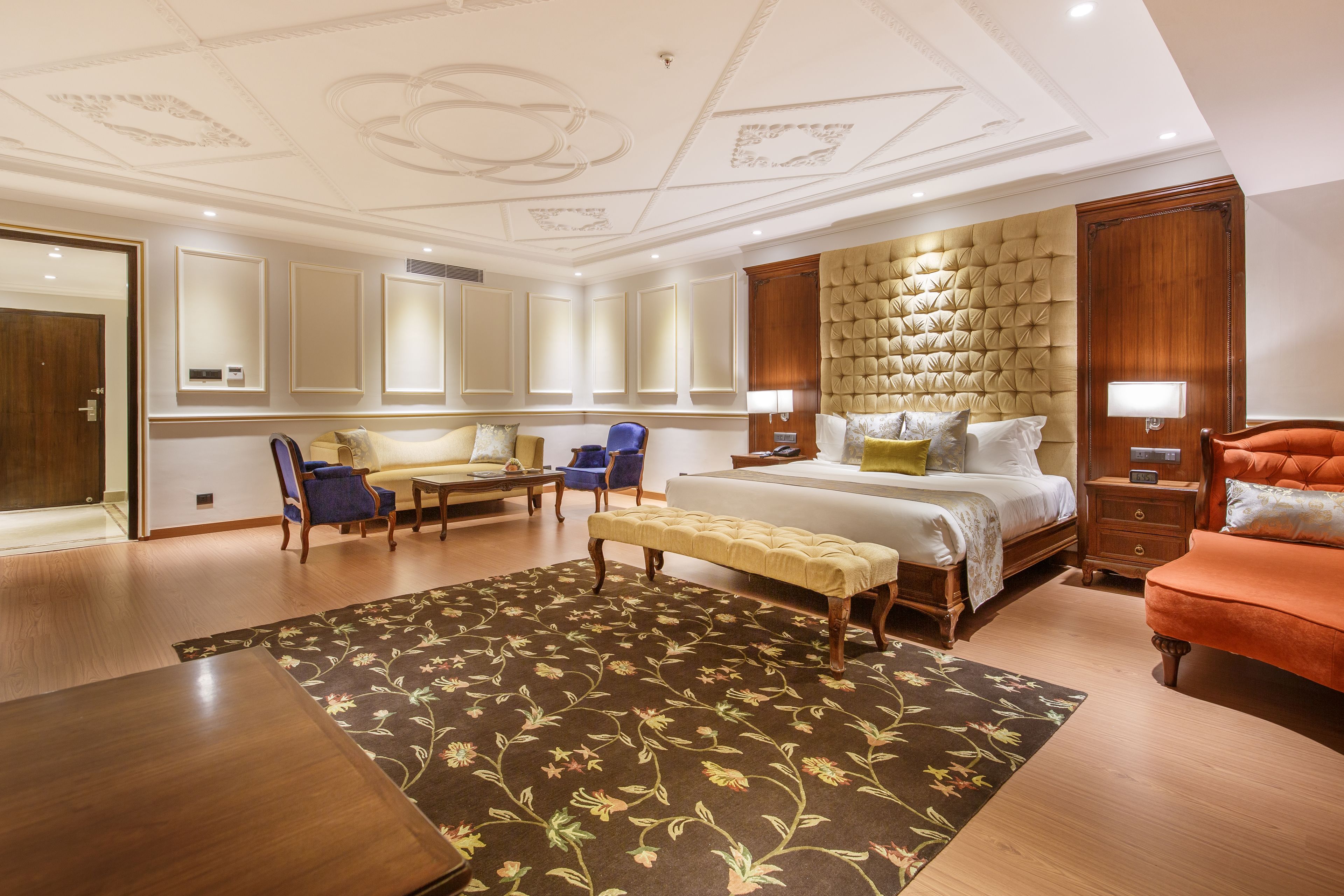 A hotel room with a large bed, a sitting area with chairs, a bench, a wooden nightstand, and a large mirror.
