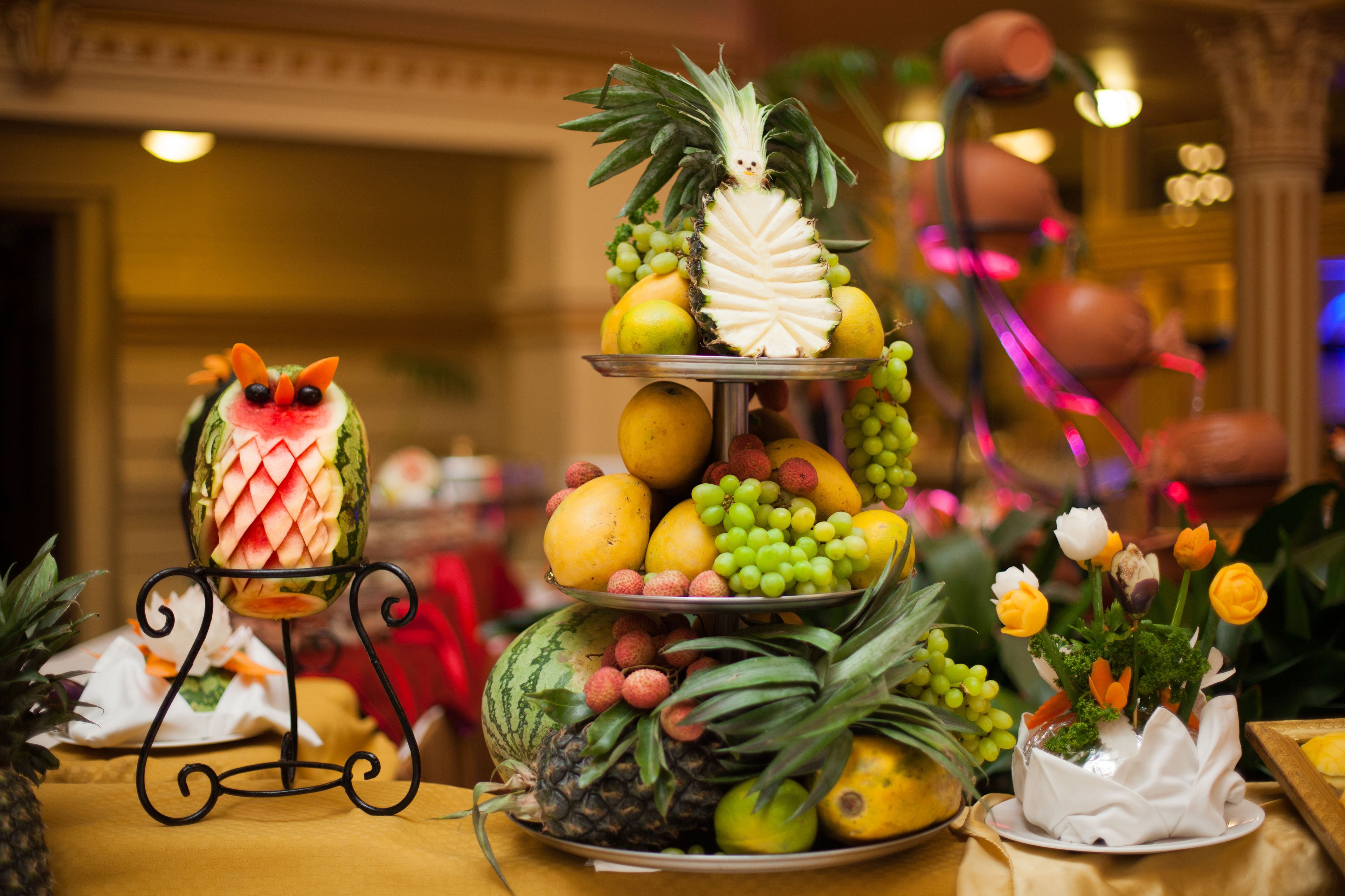 A fruit display with a watermelon carved into a fish, a pineapple, and various fruits on tiered stands.