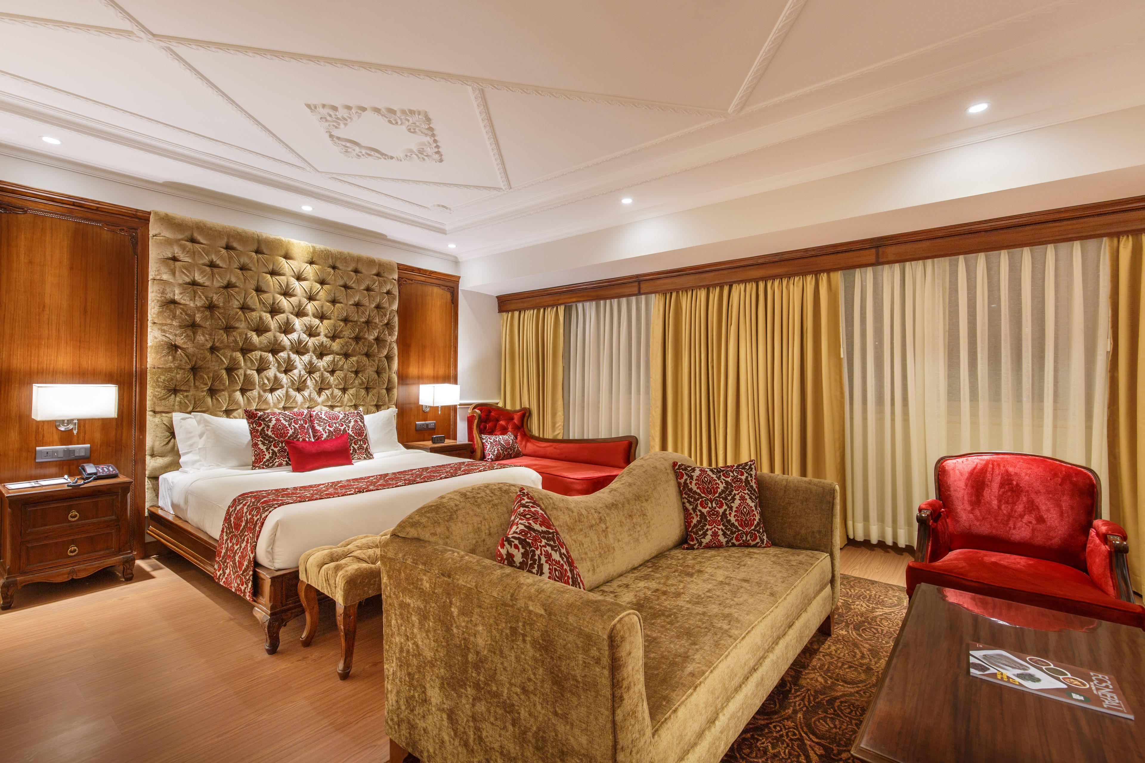 A hotel room with a large bed, a sofa, a red armchair, a wooden nightstand, and a wooden headboard.