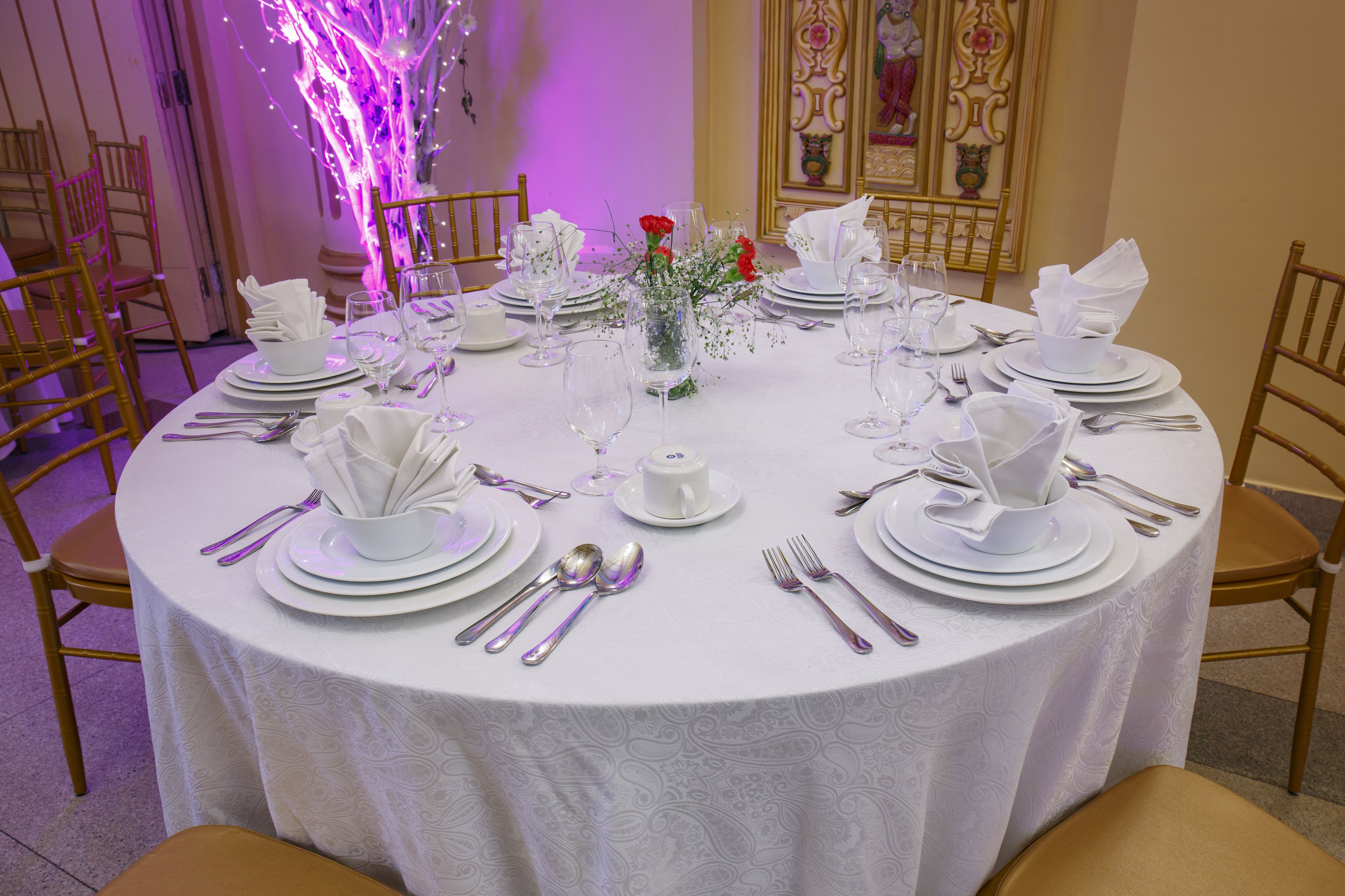 A round dining table set for a formal event with white tablecloths, silverware, and a centerpiece with red flowers.