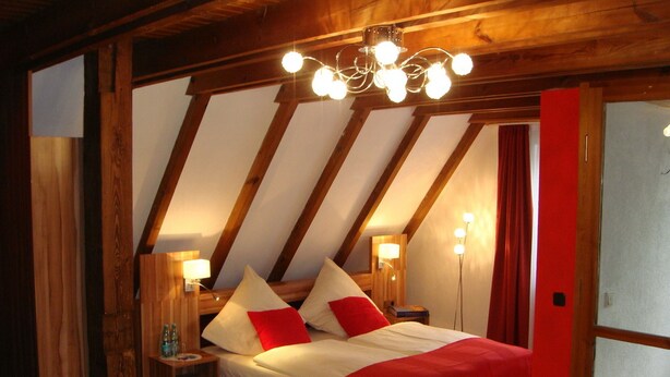 Une chambre avec un plafond en pente, des poutres en bois, un lit avec une literie rouge et blanche, des lampes de chevet et un lustre.