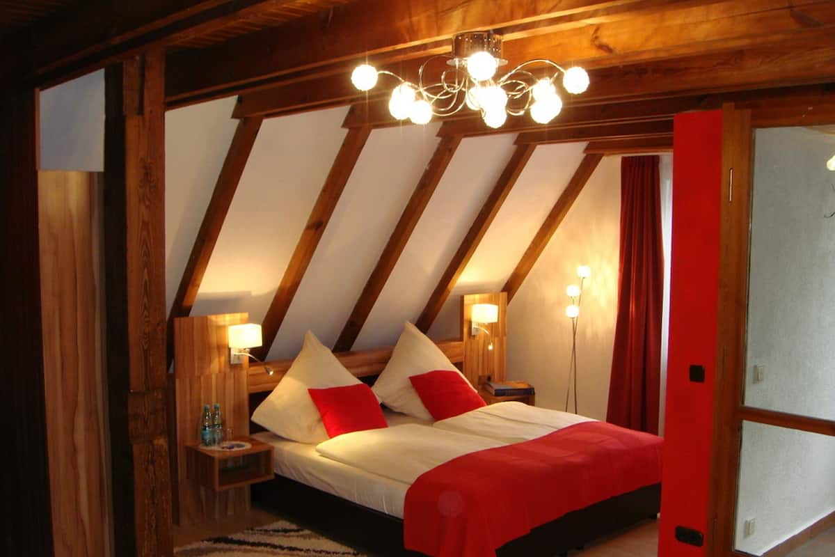 A bedroom with a sloped ceiling, wooden beams, a bed with red and white bedding, bedside lamps, and a chandelier.