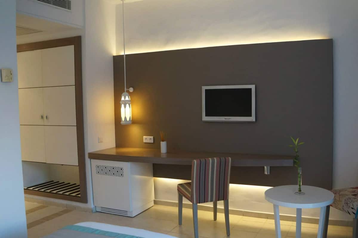 A modern, compact living space with a kitchenette, a flat-screen TV mounted on the wall, a small round table, and a striped chair.