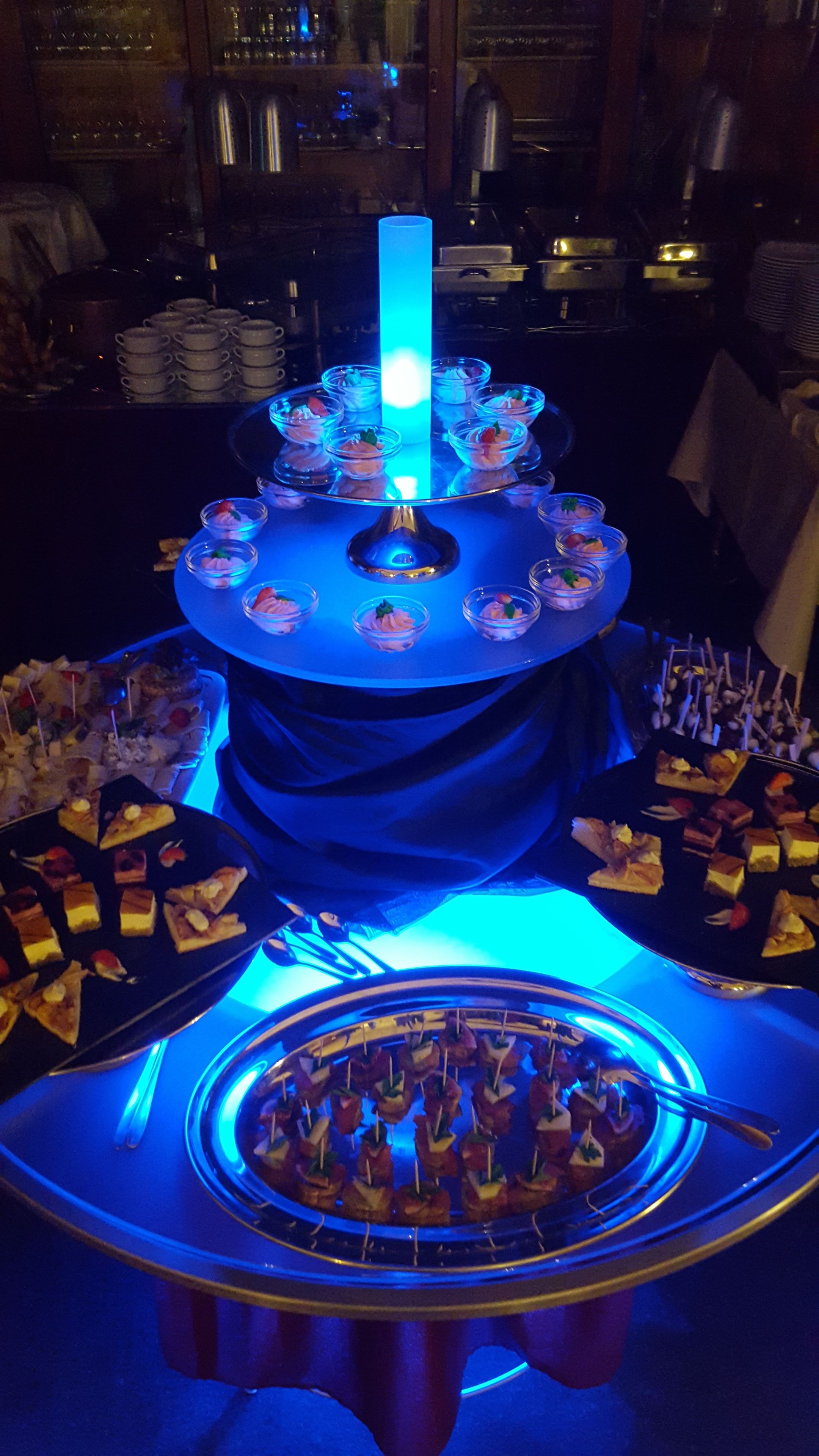 Eine mehrstufige Dessertvitrine mit blauem Licht in der Mitte, umgeben von verschiedenen Speisen und einer kreisförmigen Anordnung von Spießen am Boden.