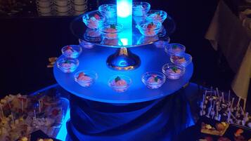 Eine mehrstufige Dessertvitrine mit blauem Licht in der Mitte, umgeben von verschiedenen Speisen und einer kreisförmigen Anordnung von Spießen am Boden.