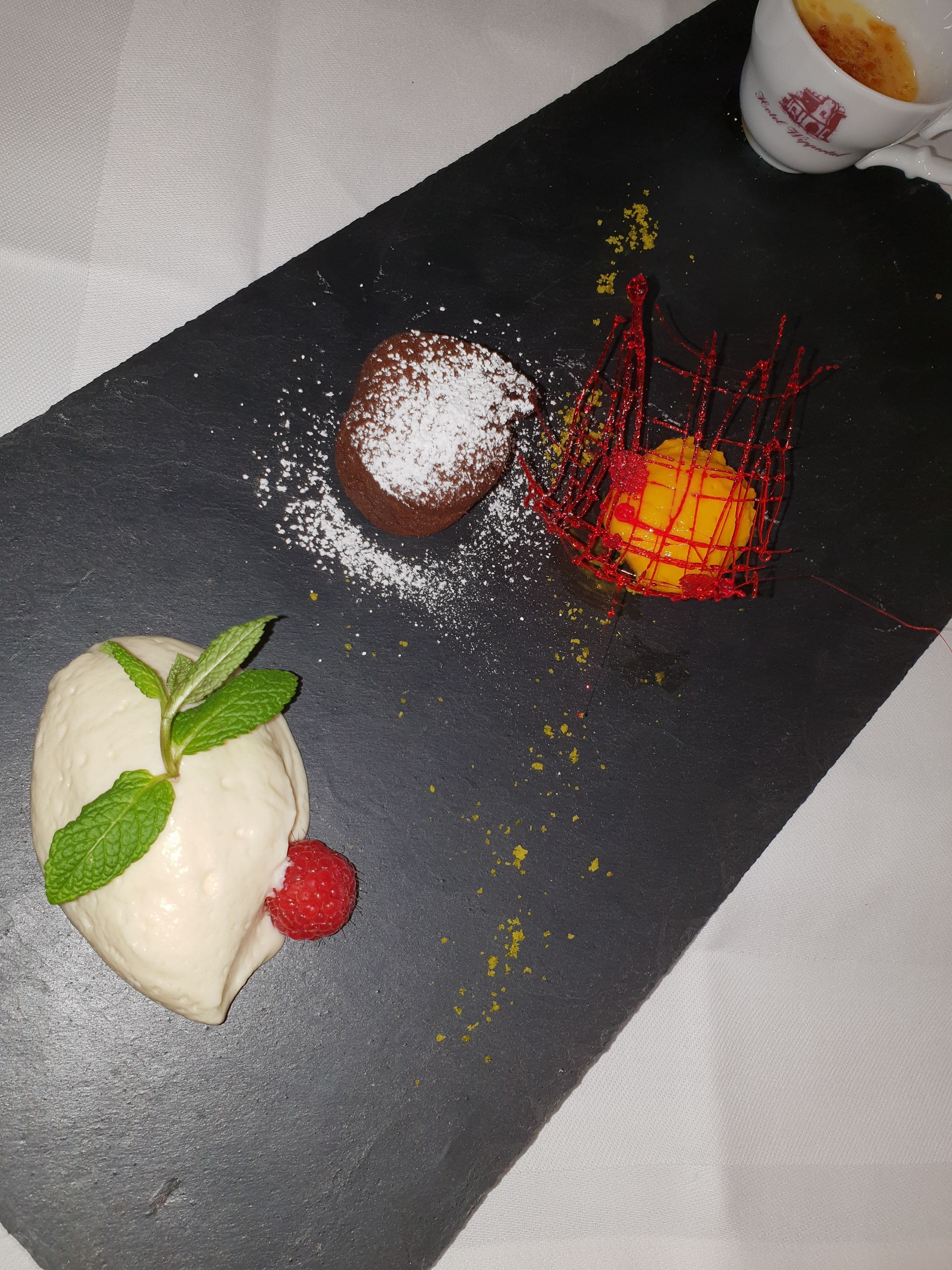 Un plat de postres amb mus de xocolata, una mango en una xarxa vermella i un dolç blanc amb maduixa i menta sobre una llosa negra.