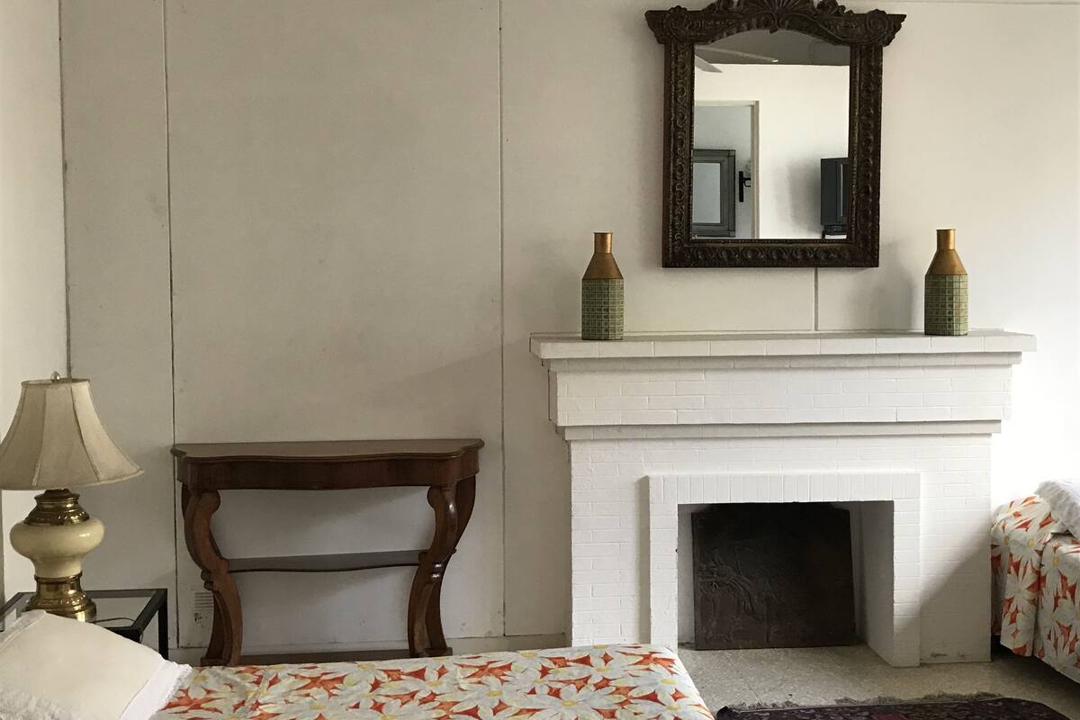 Ein Schlafzimmer mit Kamin aus weißem Backstein, einem Holztisch, einem Bett mit floralem Überwurf und zwei dekorativen Vasen auf dem Kaminvorsatz.