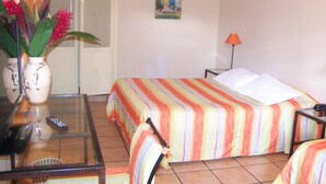 Habitación de hotel con cama, una silla, una mesa, un florero con flores, un cuadro en la pared y una lámpara.