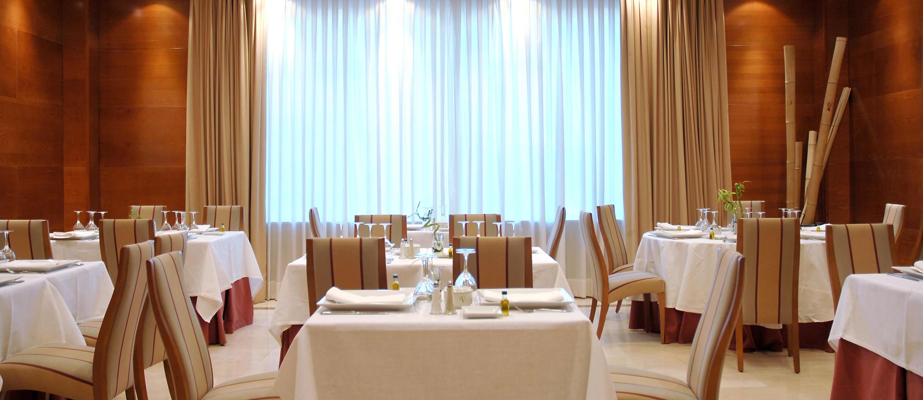 A dining area with tables set for service, large windows with curtains, and wooden paneling on the walls.