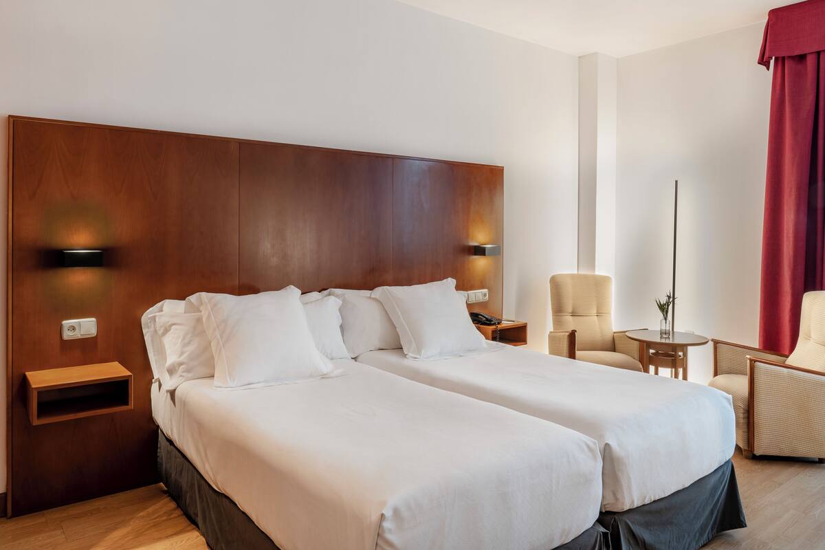 A hotel room with a large wooden headboard, two beds with white linens, a small round table, a chair, and a red curtain.