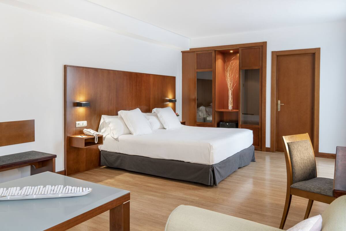 A hotel room with a large bed, a desk, a chair, a wardrobe, and a nightstand with a phone.