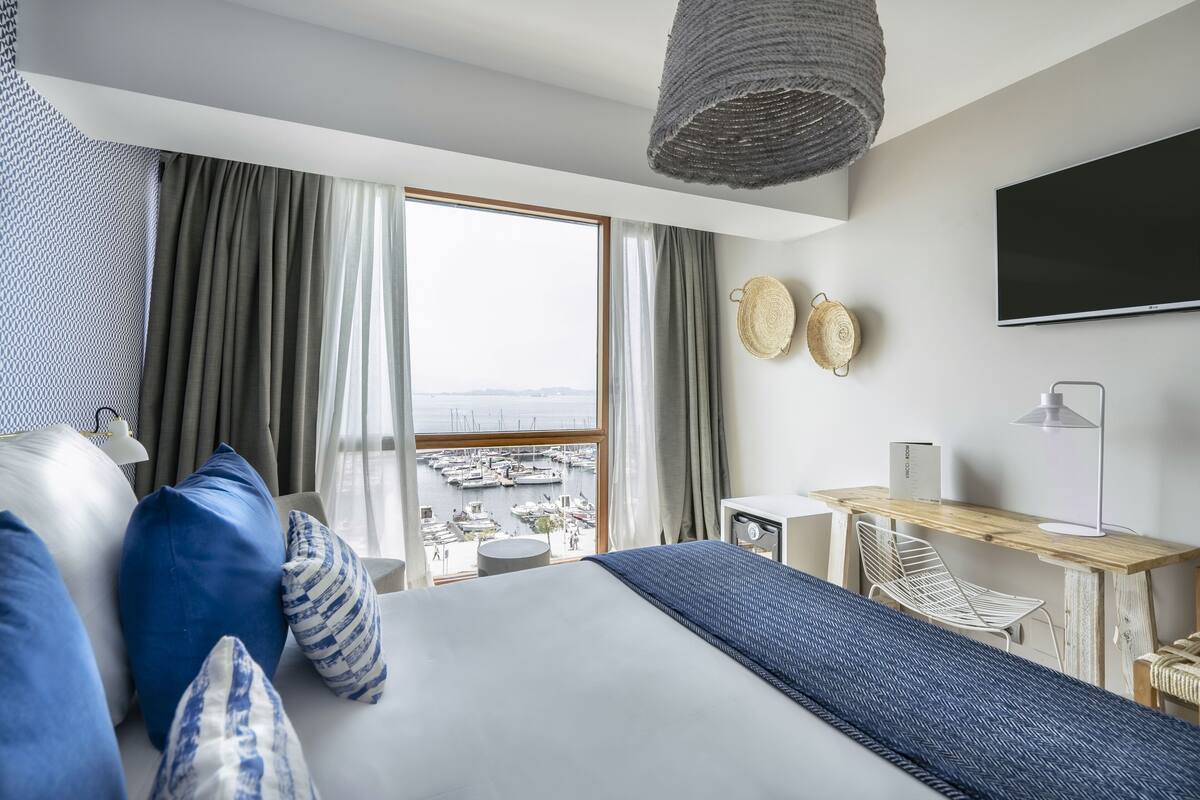 A hotel room with a large bed, a desk, a chair, a TV, and a view of a marina.