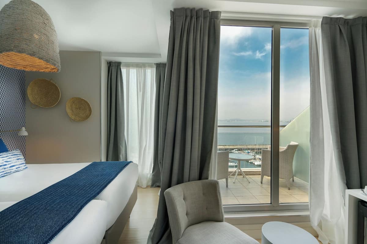 A hotel room with a bed, a chair, a small table, and a view of the sea through large windows.