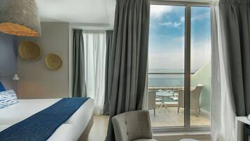 Een hotelkamer met een bed, een stoel, een tafeltje en uitzicht op zee door grote ramen.