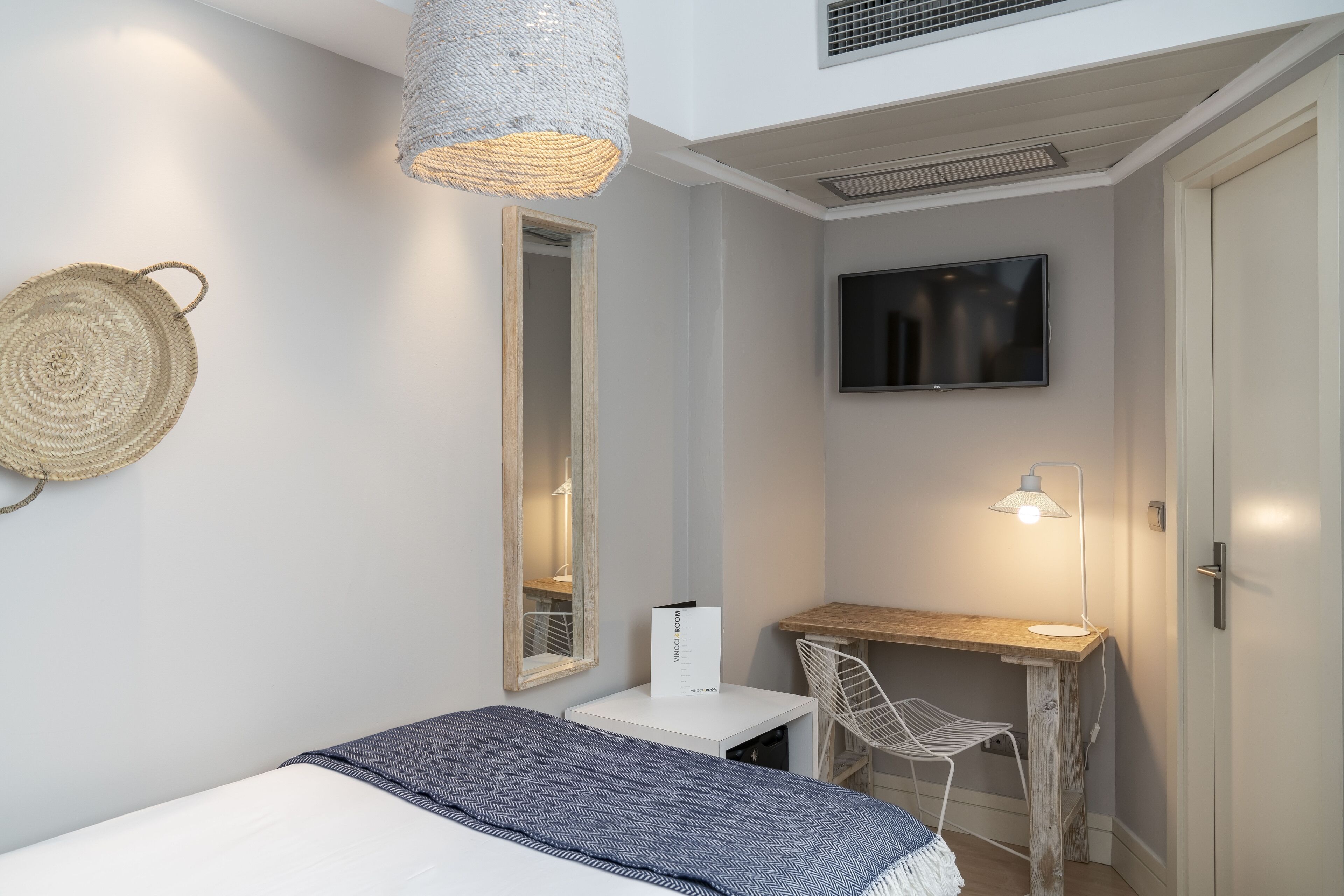 A compact bedroom with a bed, a desk, a chair, a TV, and a woven wall hanging.