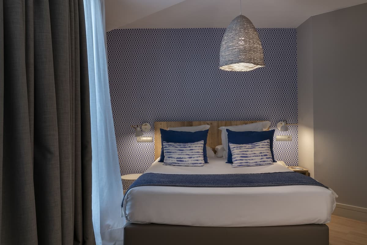 A neatly made bed with a blue and white striped pillow, a hanging light fixture, and patterned wallpaper.