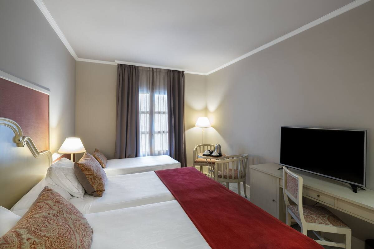 A hotel room with two beds, a desk with a TV, a lamp, and a window with curtains.