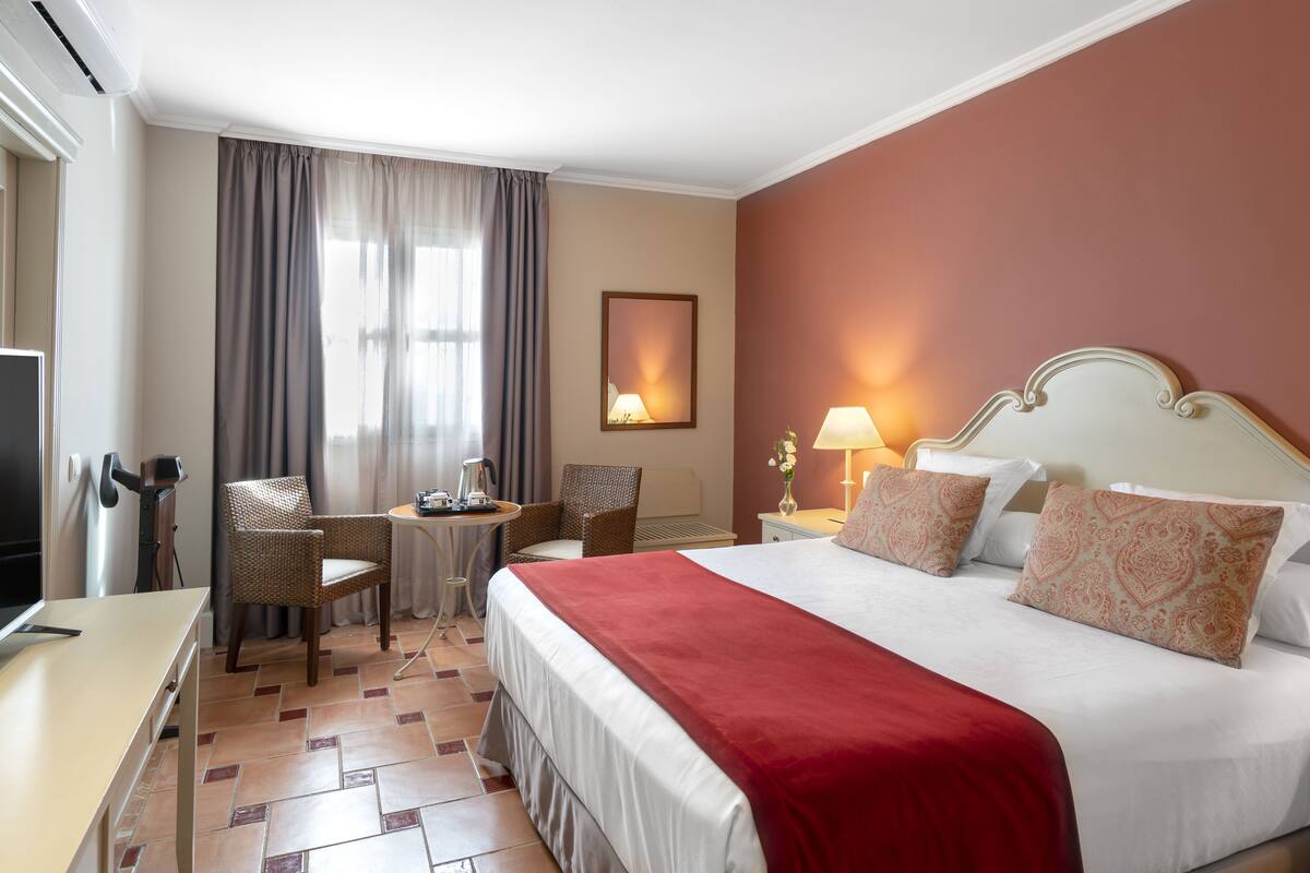 A hotel room with a large bed, a desk, a chair, a small table, and a lamp.