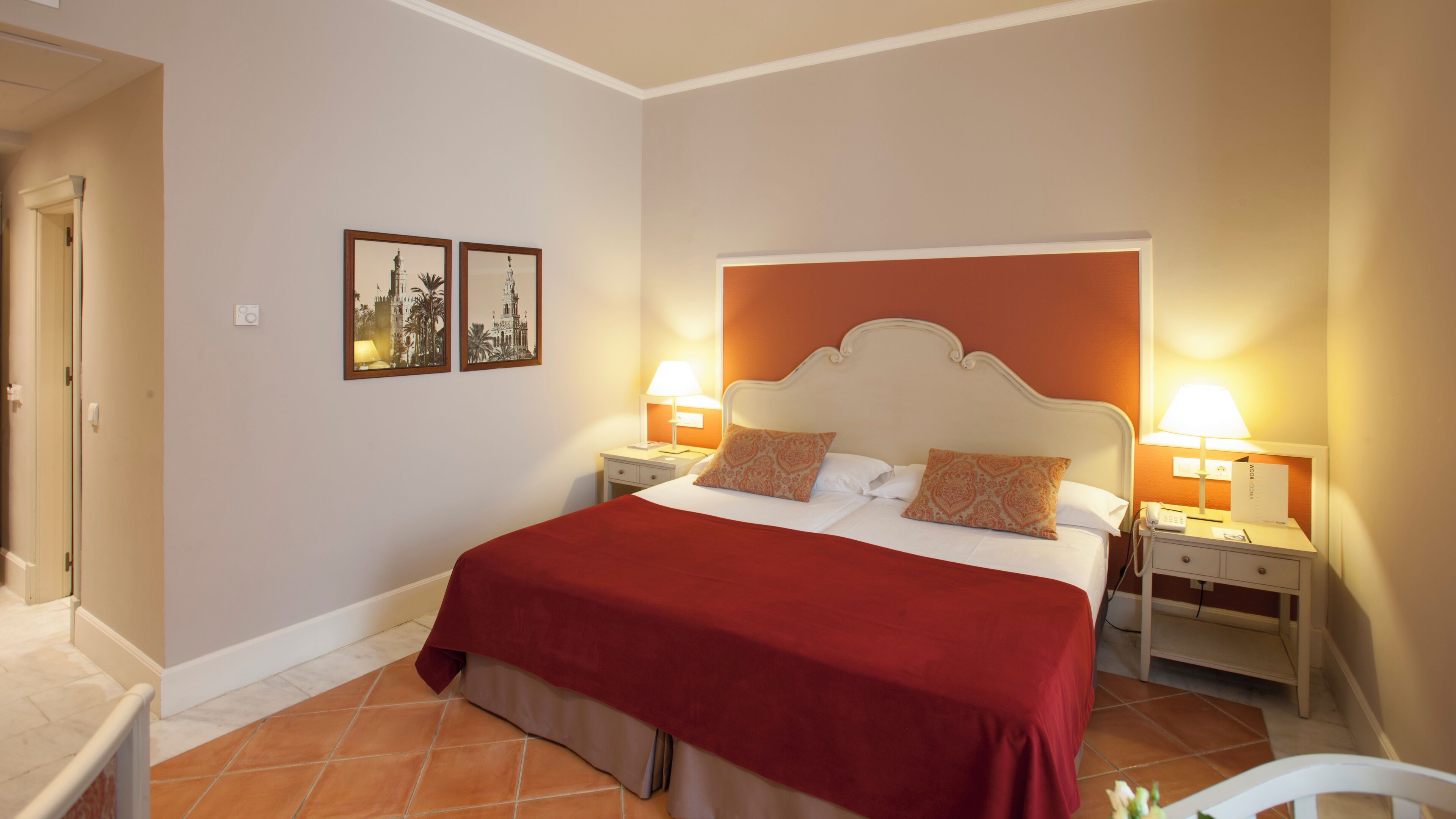 A hotel room with a large bed, two bedside tables with lamps, a red bedspread, and two framed pictures on the wall.