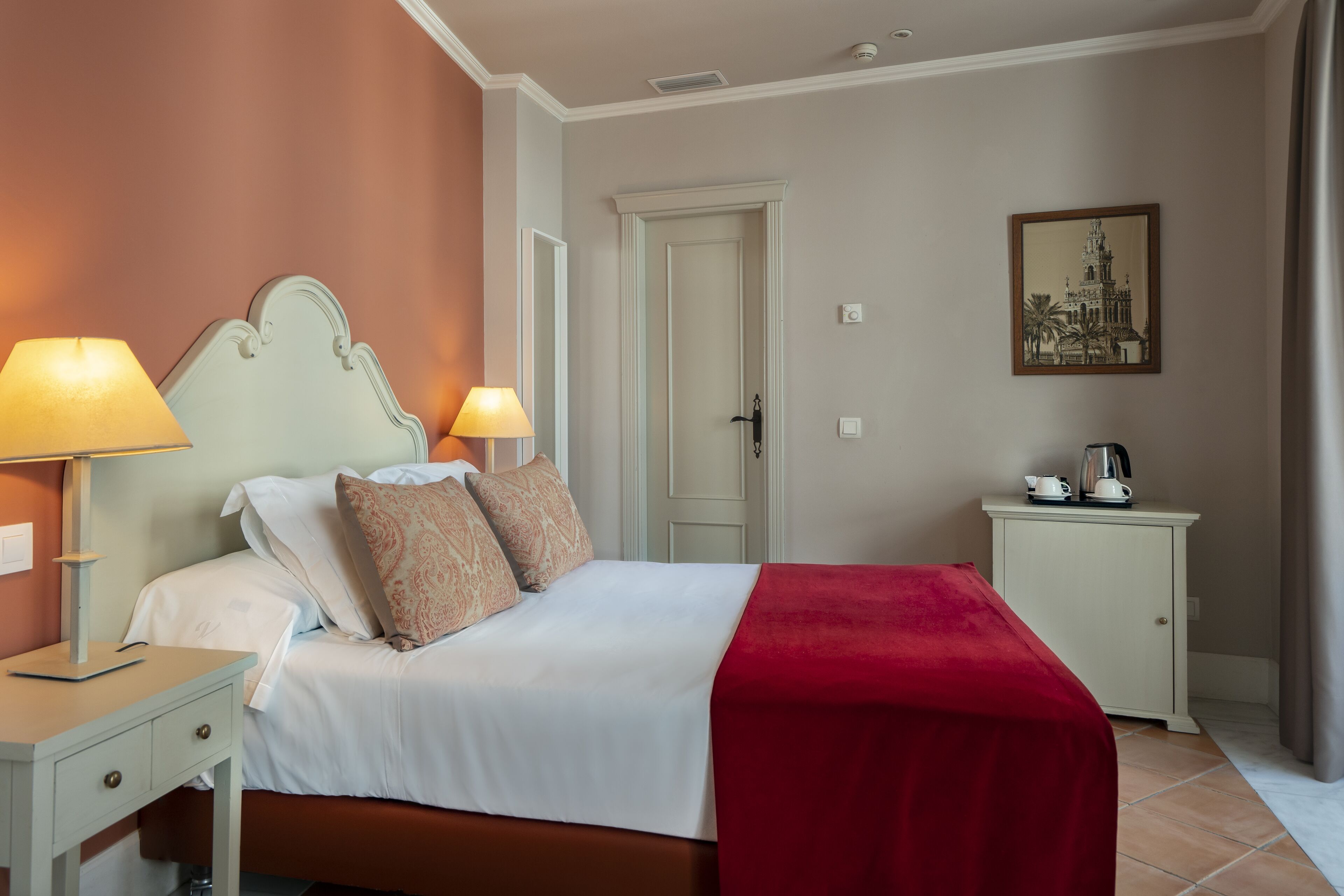 Un dormitorio ordenado con cama, lámparas de noche, una manta roja, mesita de noche, armario y un cuadro en la pared.