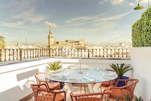 Une terrasse sur le toit, avec une table ronde dressée pour le dîner, entourée de chaises en osier, et offrant une vue sur un paysage urbain historique dominé par un imposant beffroi.