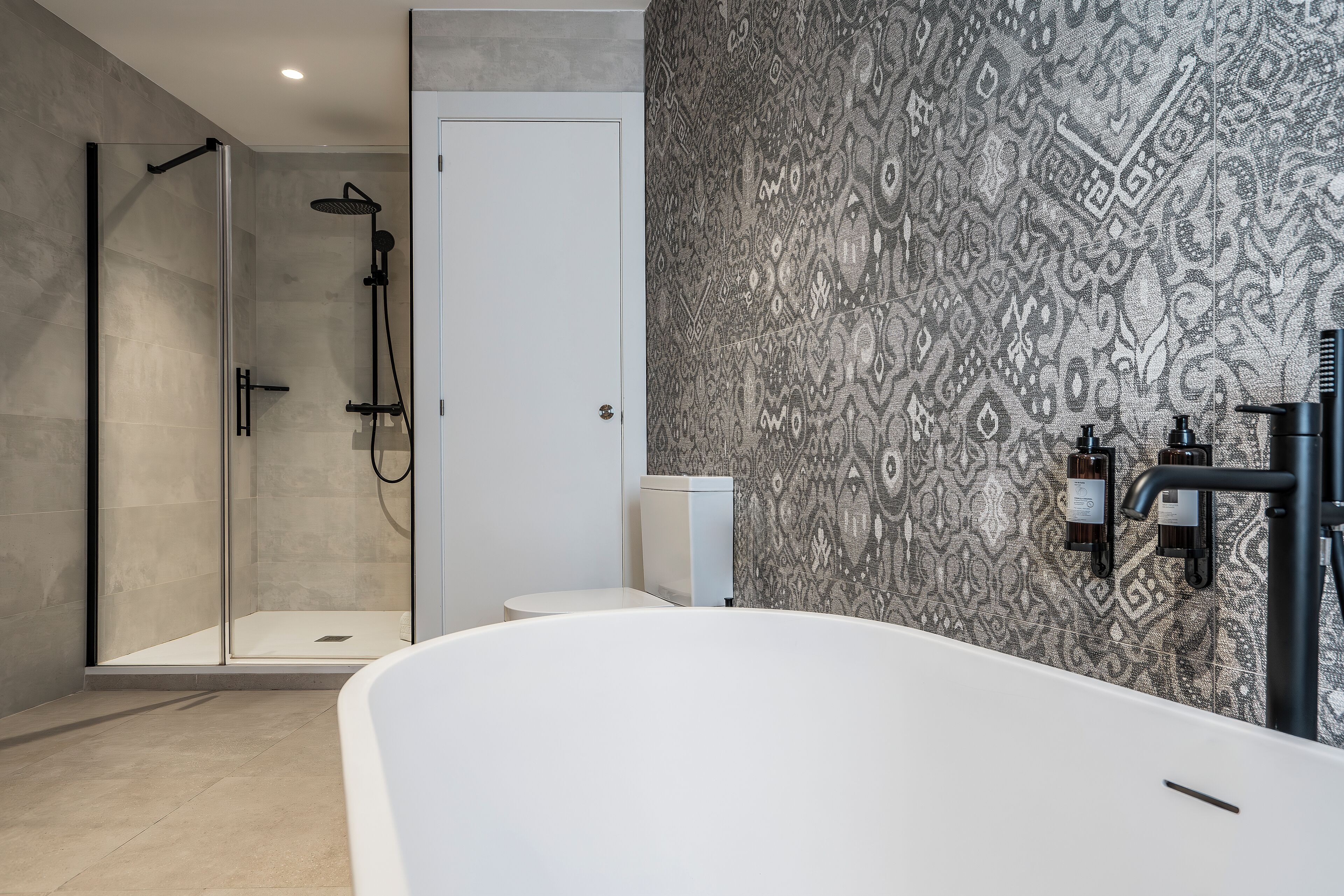 A modern bathroom with a glass-enclosed shower, a white bathtub, and patterned wall tiles.