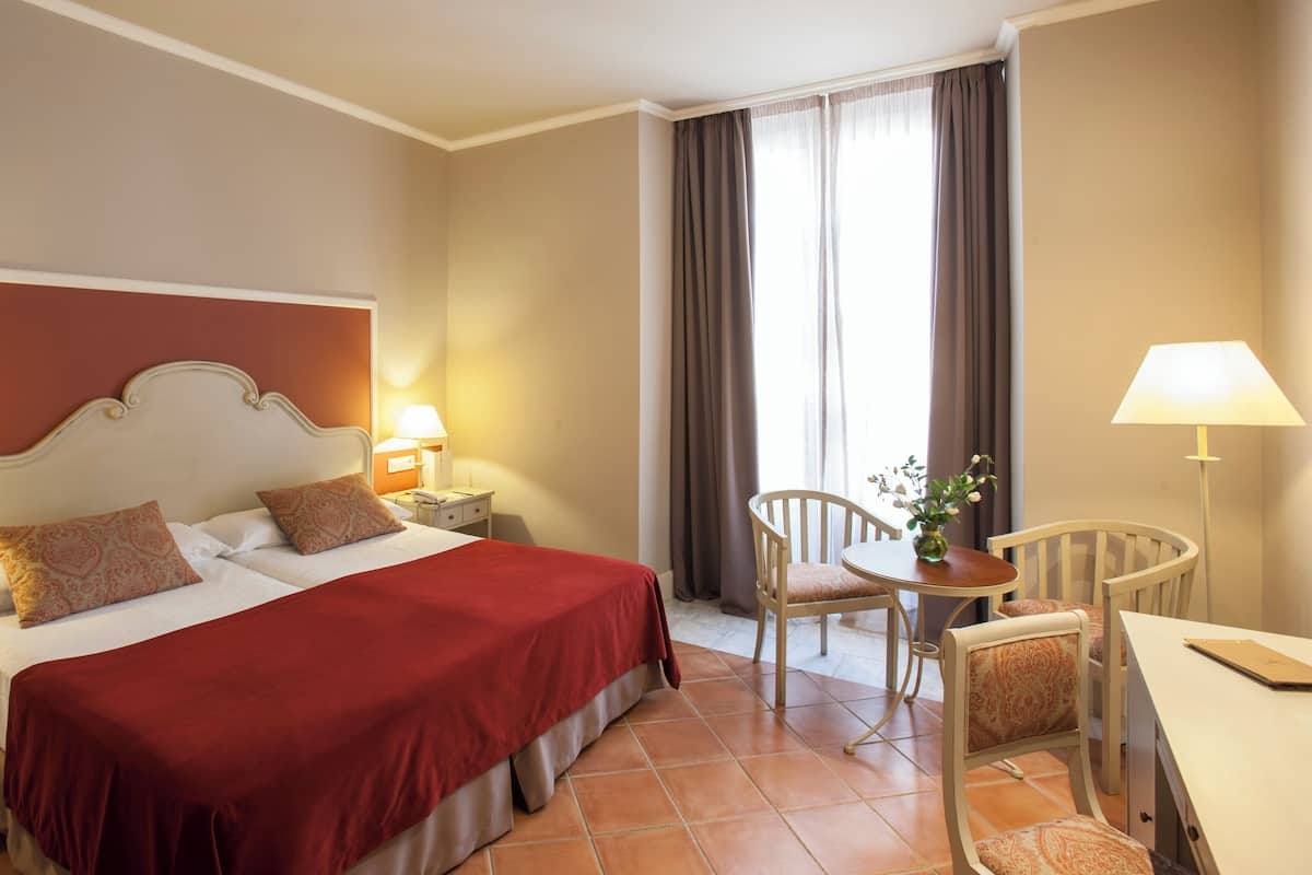 A hotel room with a bed, bedside tables, a desk, a chair, and a window with curtains.