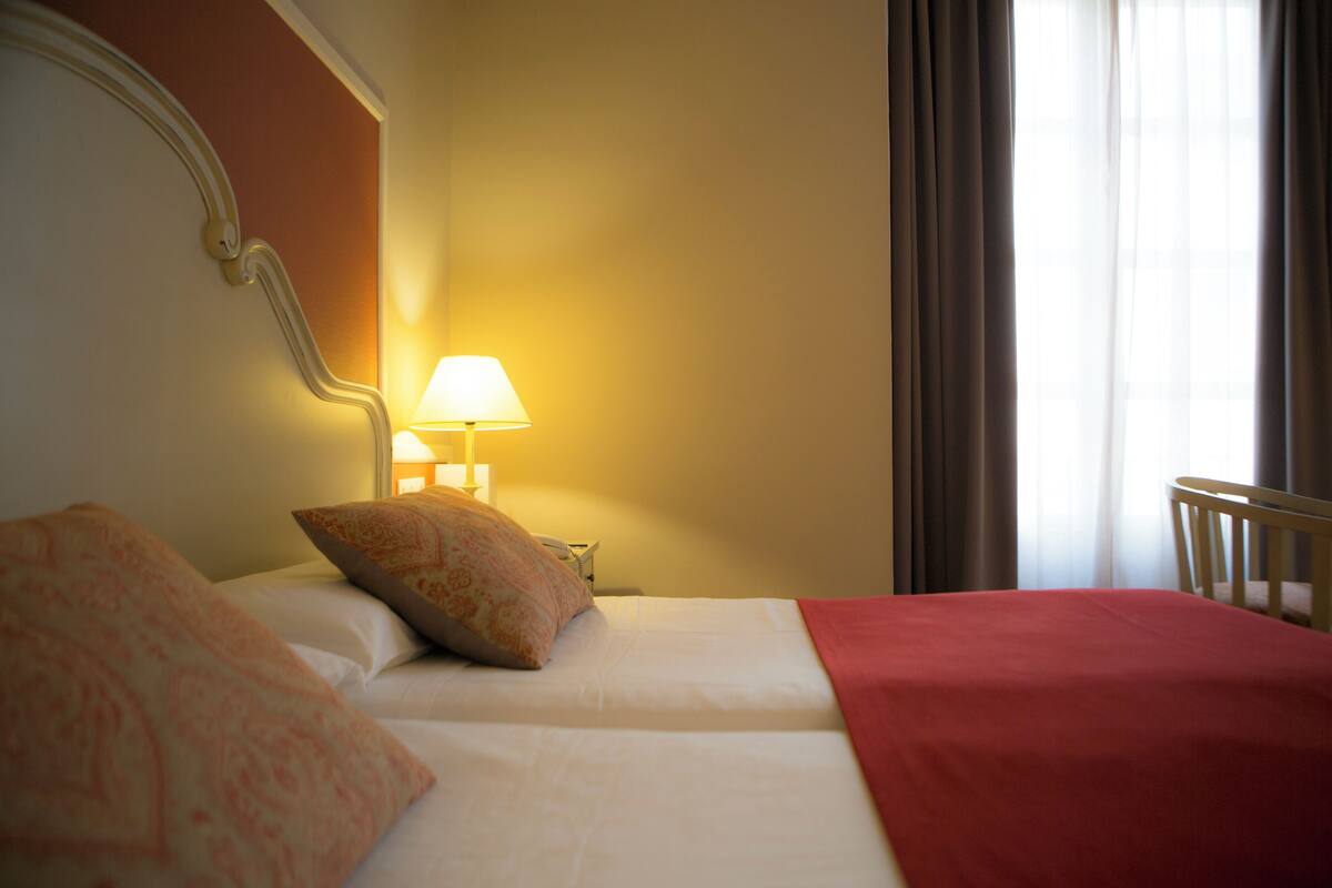 A hotel room with a bed, a lamp, a chair, and a window with curtains.
