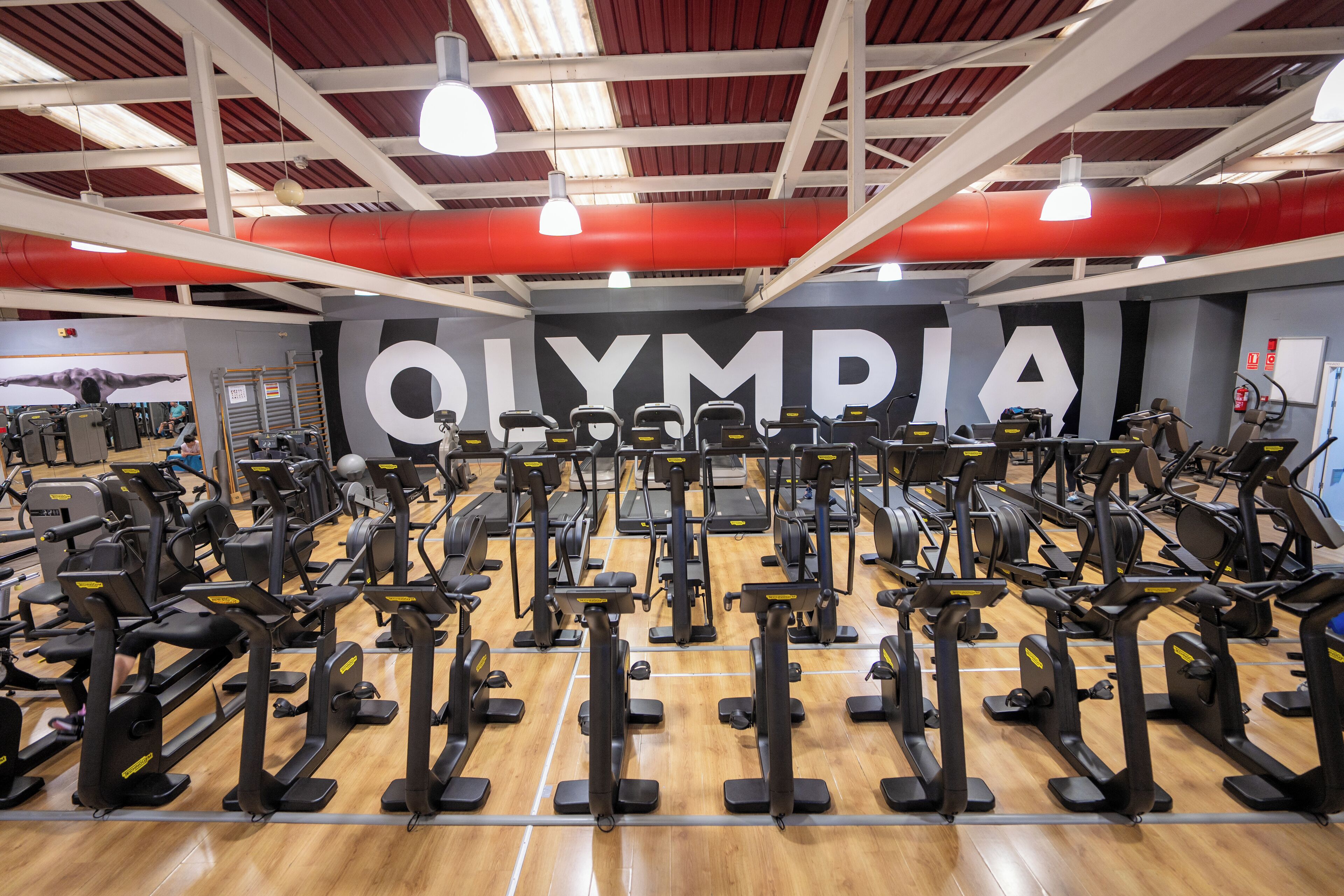 Une salle de sport équipée de nombreux appareils d’exercice, un grand panneau « OLYMPIA » et un plafond haut avec des poutres rouges et des luminaires suspendus.