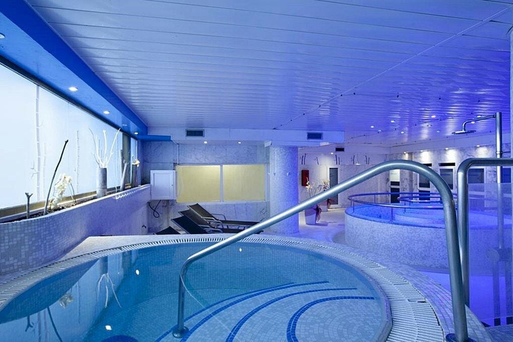 Una piscina interior con borde de azulejos azules, barandilla metálica y barrera de vidrio.