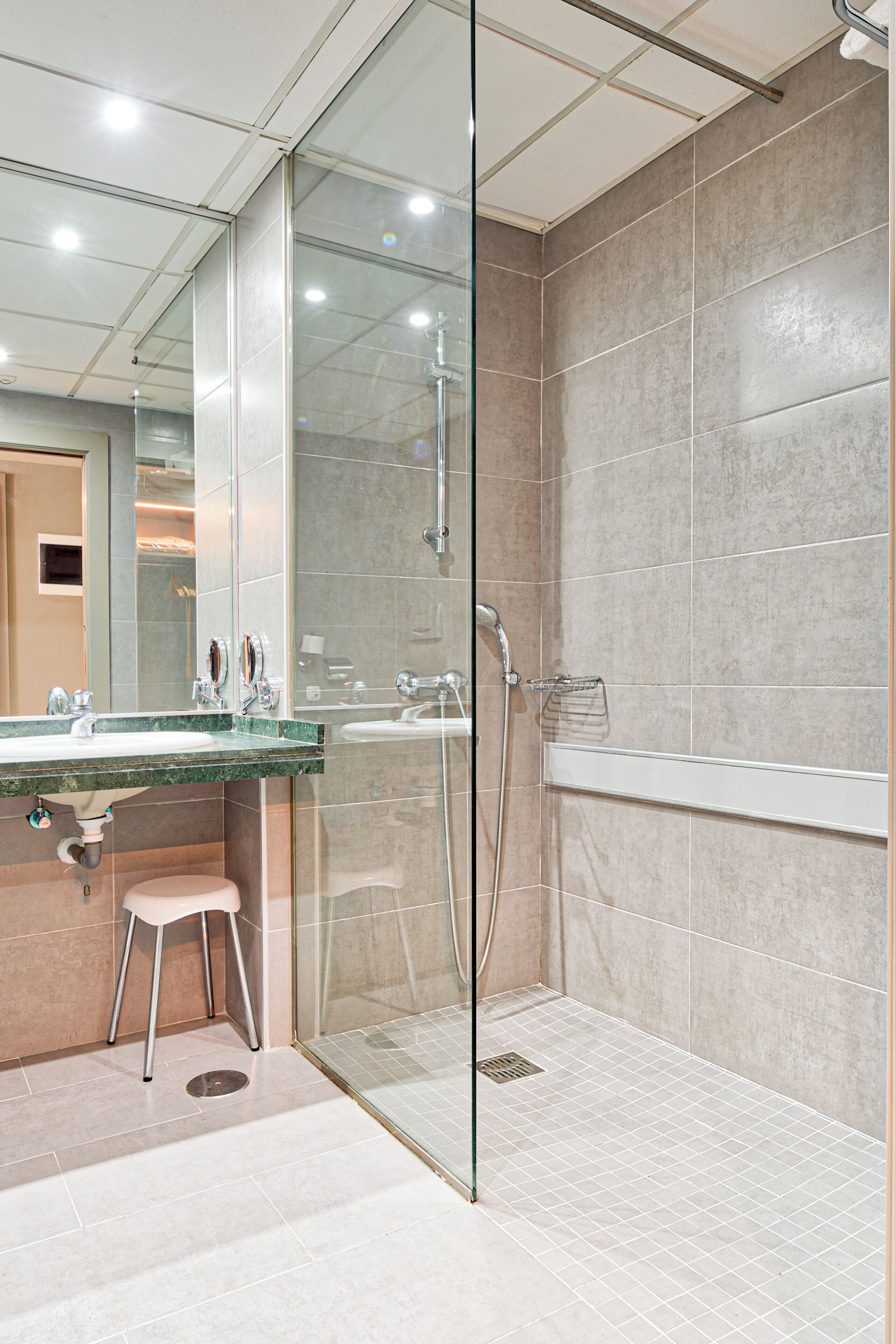 Un baño moderno con ducha envuelta en cristal, un lavamanos con encimera de mármol y un taburete pequeño.