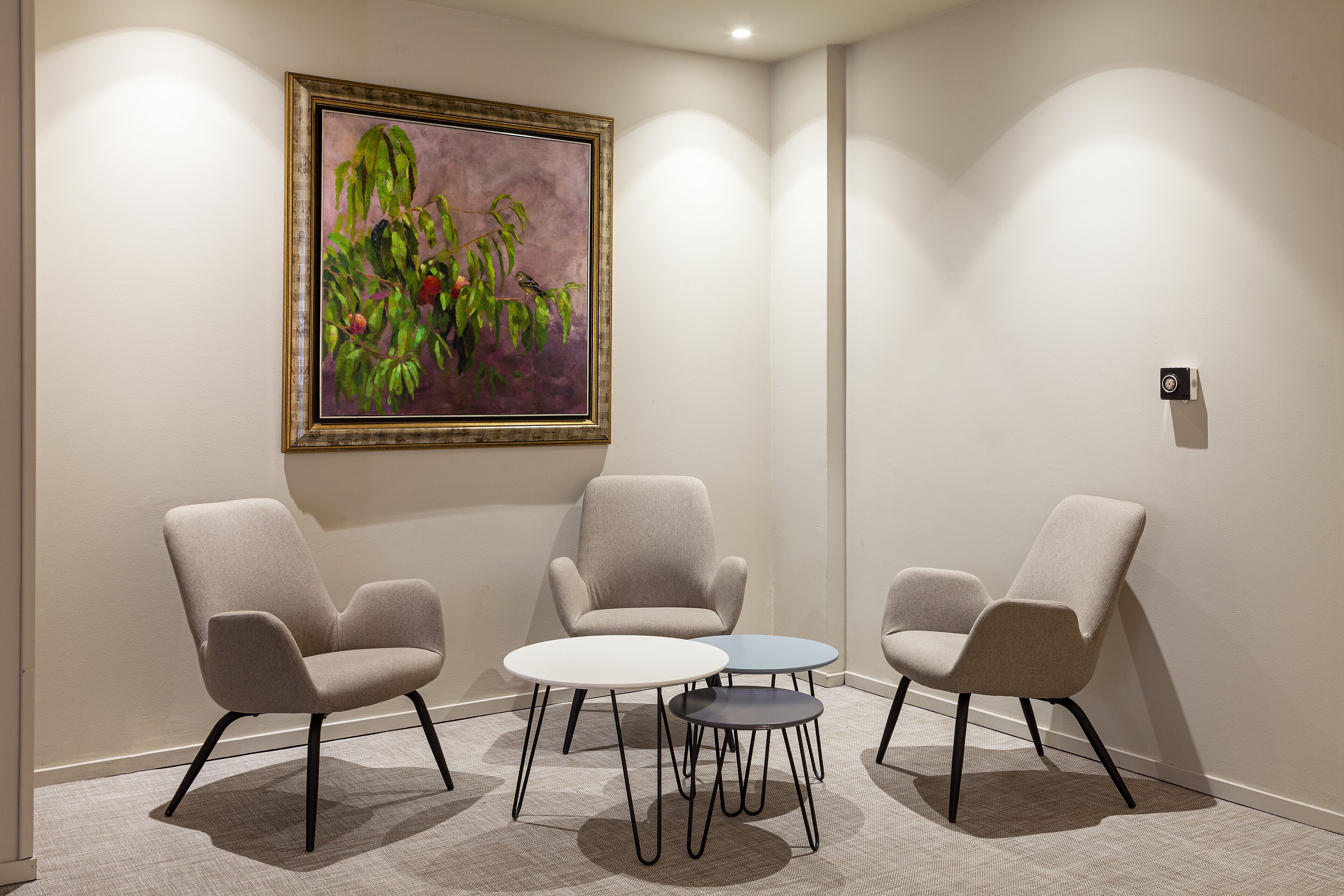 Una stanza con tre sedie e due tavoli, uno rotondo e uno ovale, e un quadro appeso al muro.