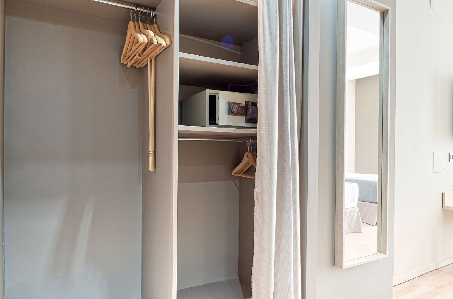 Un armario espacioso con perchas y estantes de madera, un espejo, y una cama visible a través de una puerta abierta.