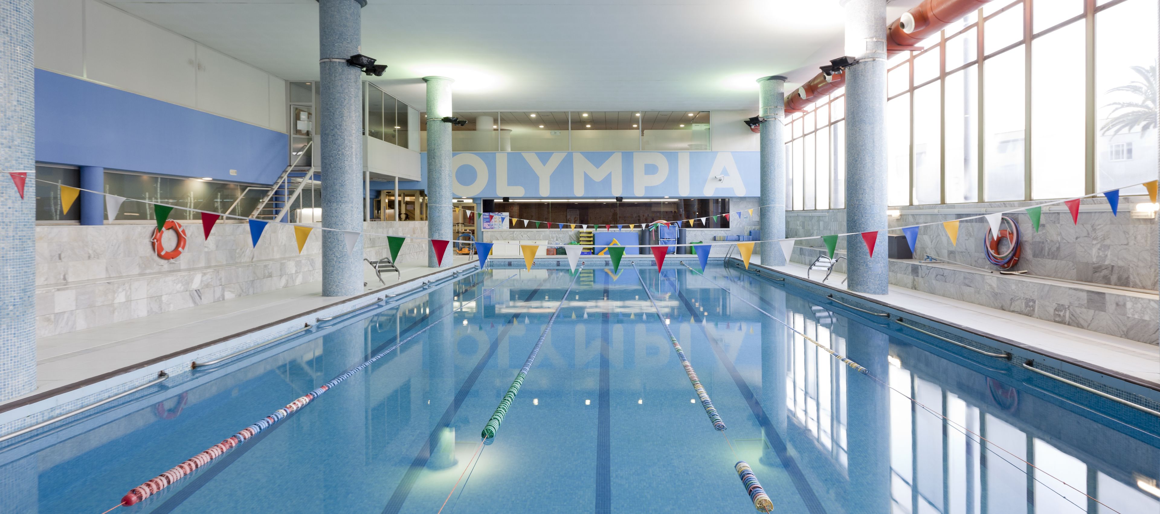 Une piscine intérieure avec des couloirs bien délimités, un grand panneau portant l’inscription « OLYMPIA », et une vue sur l’intérieur.