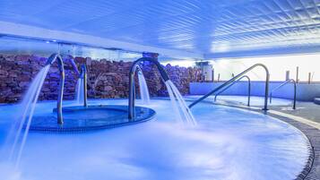 Una piscina interior moderna con múltiples chorros de agua, paredes de piedra y un techo con diseño geométrico.