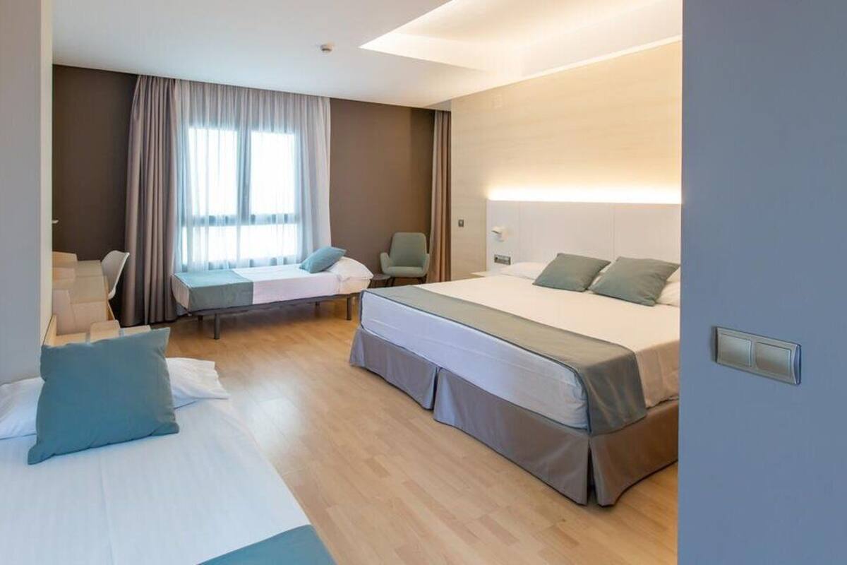 Une chambre d’hôtel avec deux lits, un bureau et une chaise. Il y a une fenêtre avec des rideaux, une table de chevet et une lampe fixée au mur.