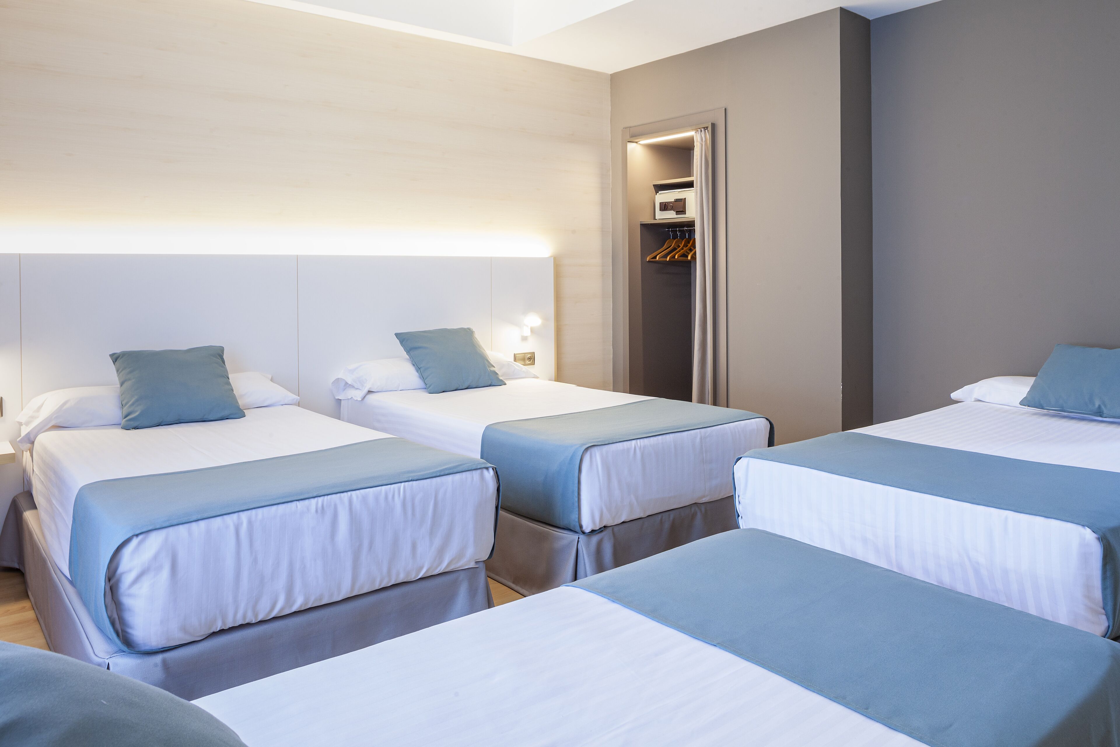 A hotel room with three single beds, each with a blue and white duvet, a bedside table with a lamp, and a built-in wardrobe with a mirror.