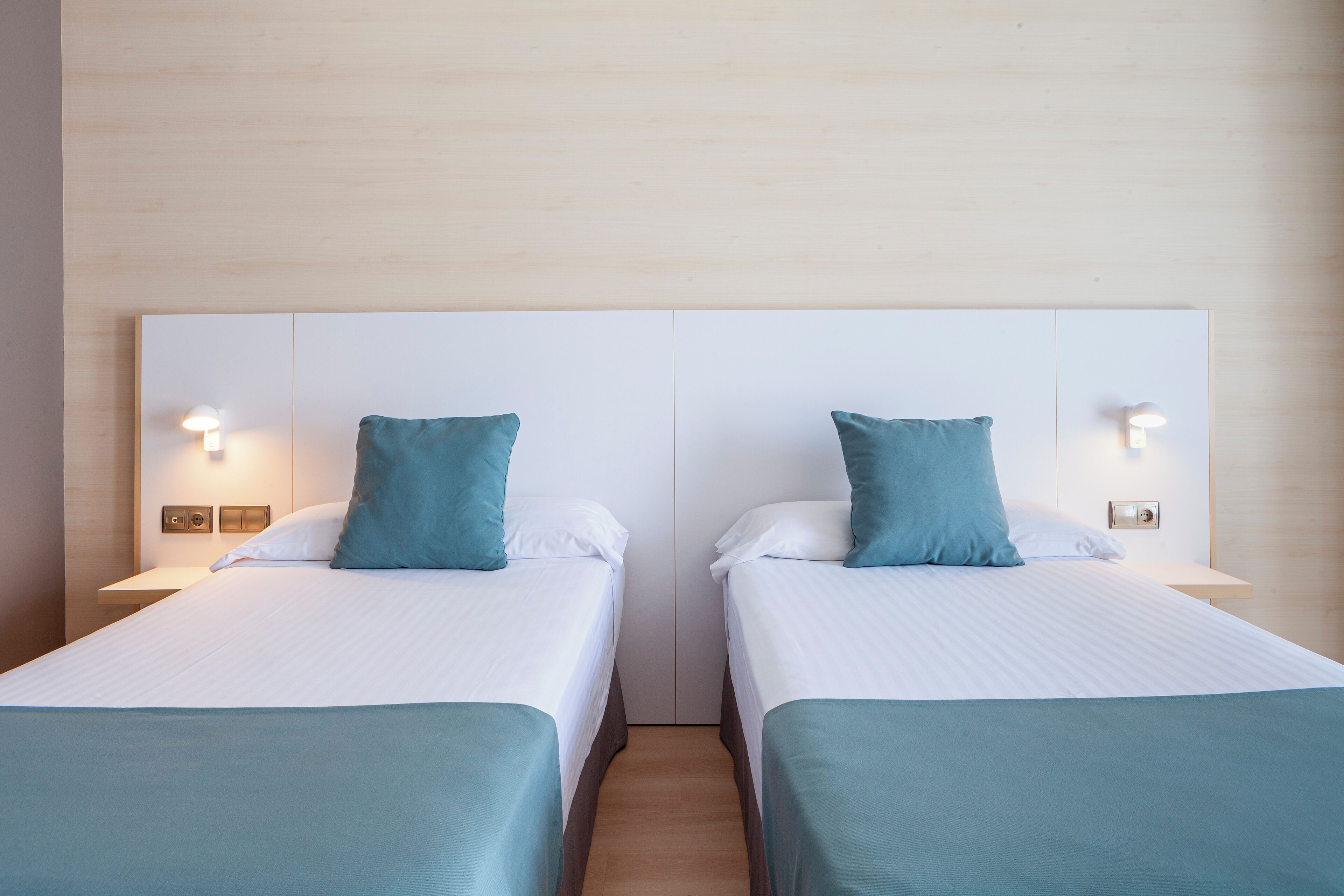 A room with two beds, each with a white headboard and a teal pillow. There are bedside tables with lamps and wall-mounted light switches.
