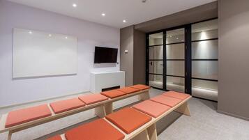 Een vergaderzaal met een groot whiteboard, een flatscreen-tv en meerdere tafels met oranje kussens.