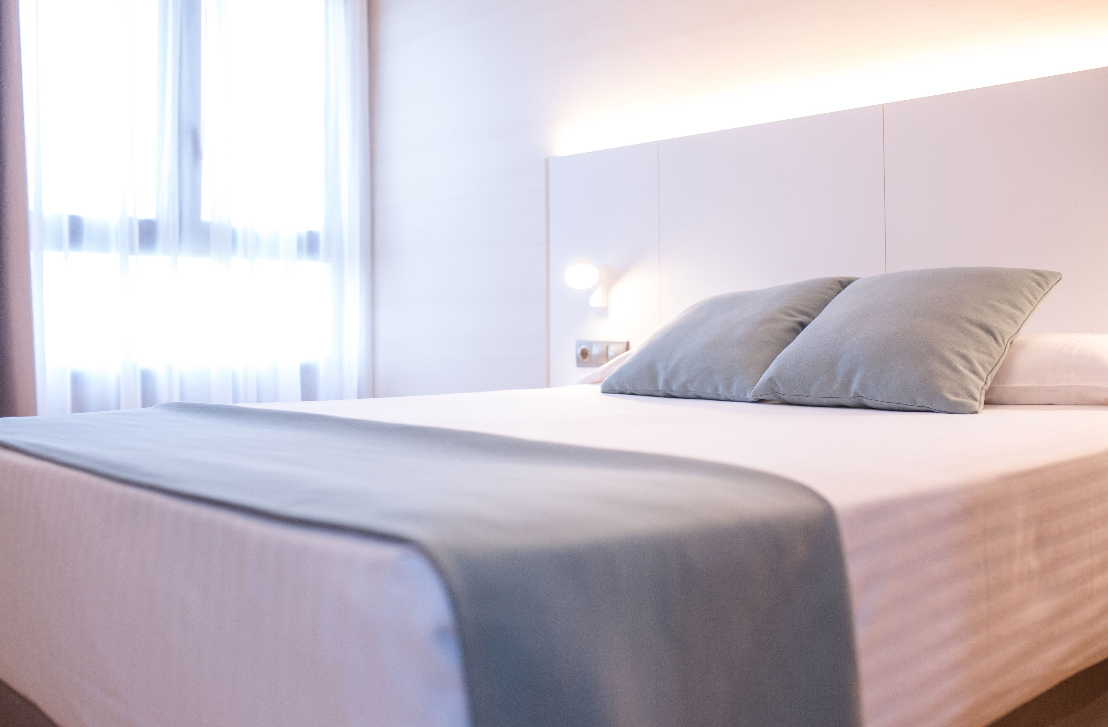 Una habitación de hotel moderna con una cama bien hecha, una lámpara de noche y un ventanal con cortinas traslúcidas.