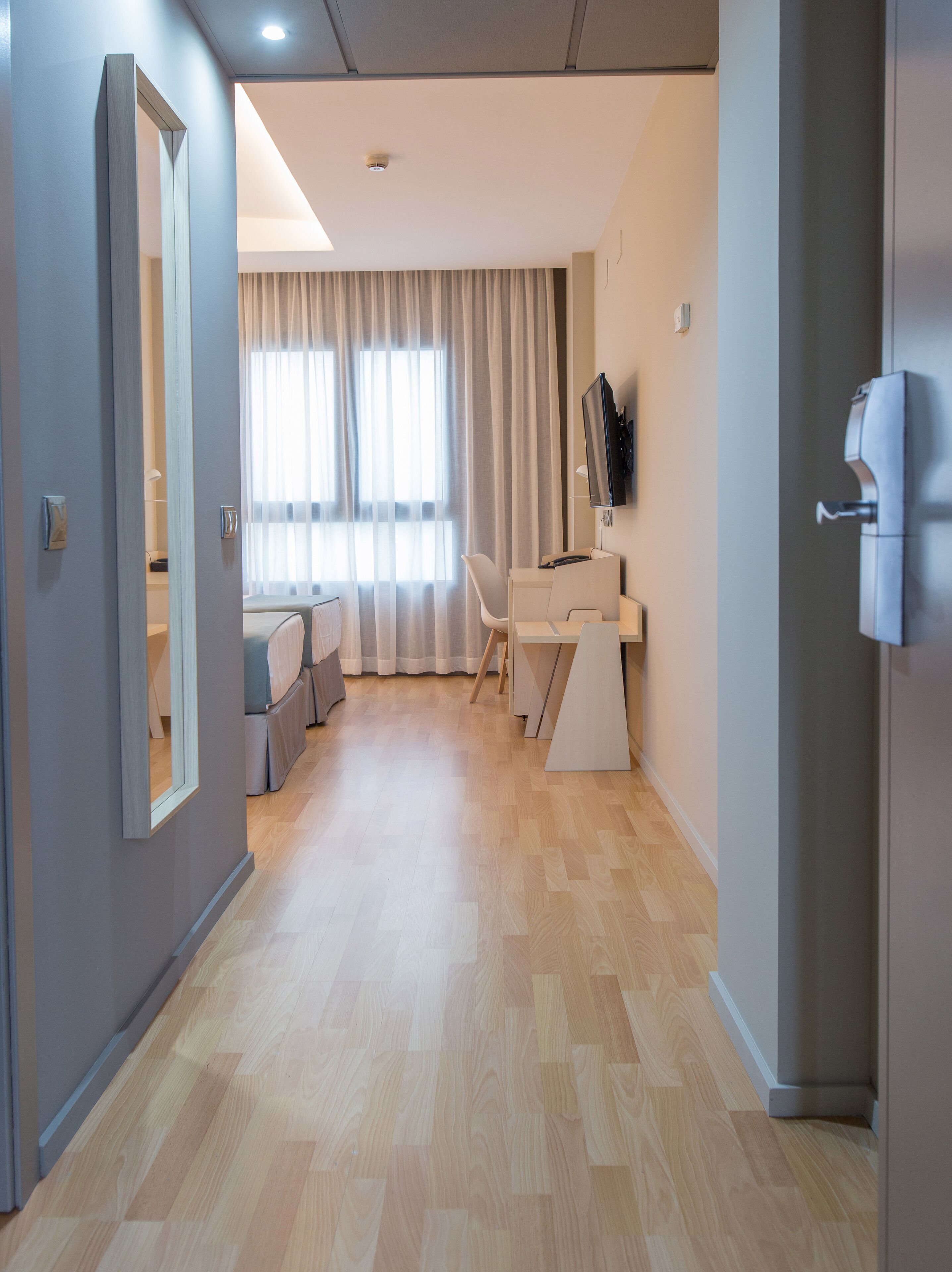 Ein modernes Hotelzimmer mit Holzboden, einem großen Spiegel, einem Schreibtisch mit Stuhl, einem Fernseher und einem Fenster mit Vorhängen.