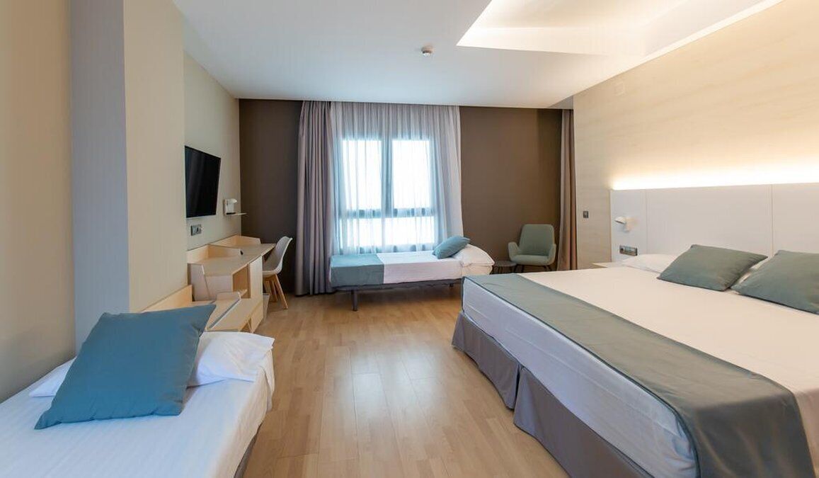 A hotel room with two single beds, a desk with a chair, a television, and a window with curtains.