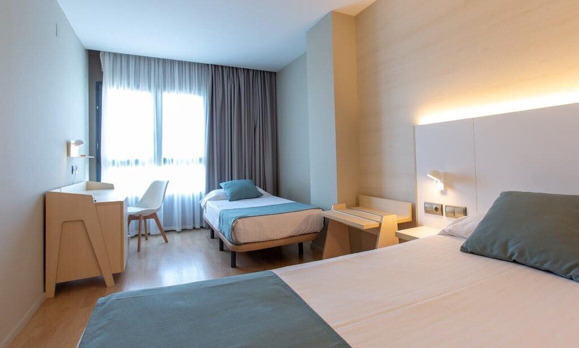 A modern hotel room with a large bed, a desk, a chair, and a small table. There is a window with curtains and a view of the outside.