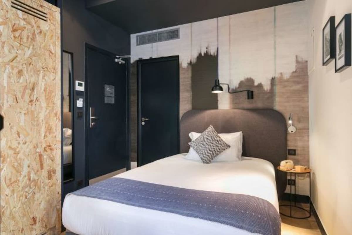 Quarto de hotel moderno com uma cama grande, cabeceira com mural de paisagem urbana, pequena mesa de cabeceira com luminária e guarda-roupa com textura de madeira.