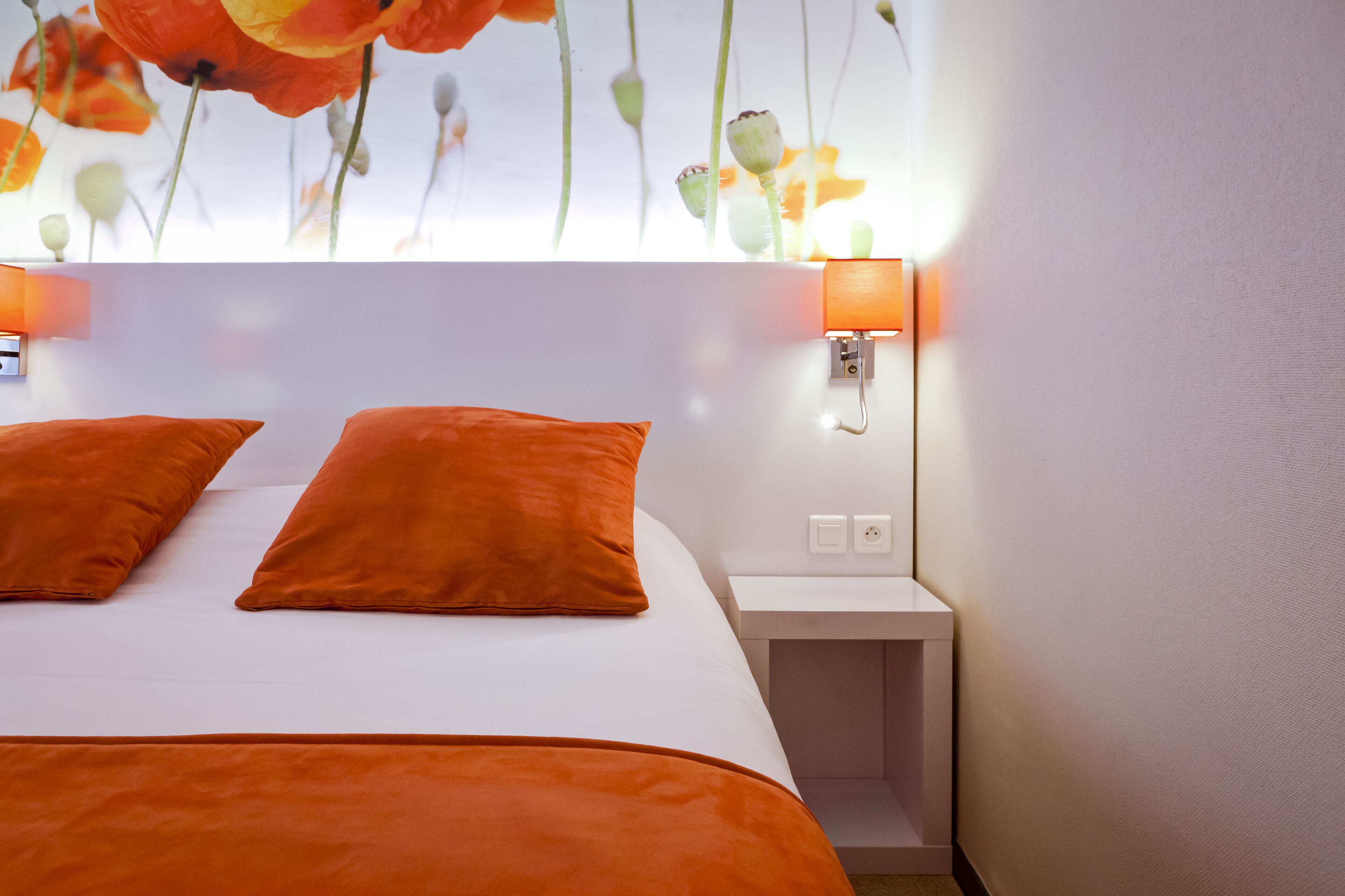 A hotel room with a bed, two orange pillows, a nightstand, and a wall-mounted lamp.