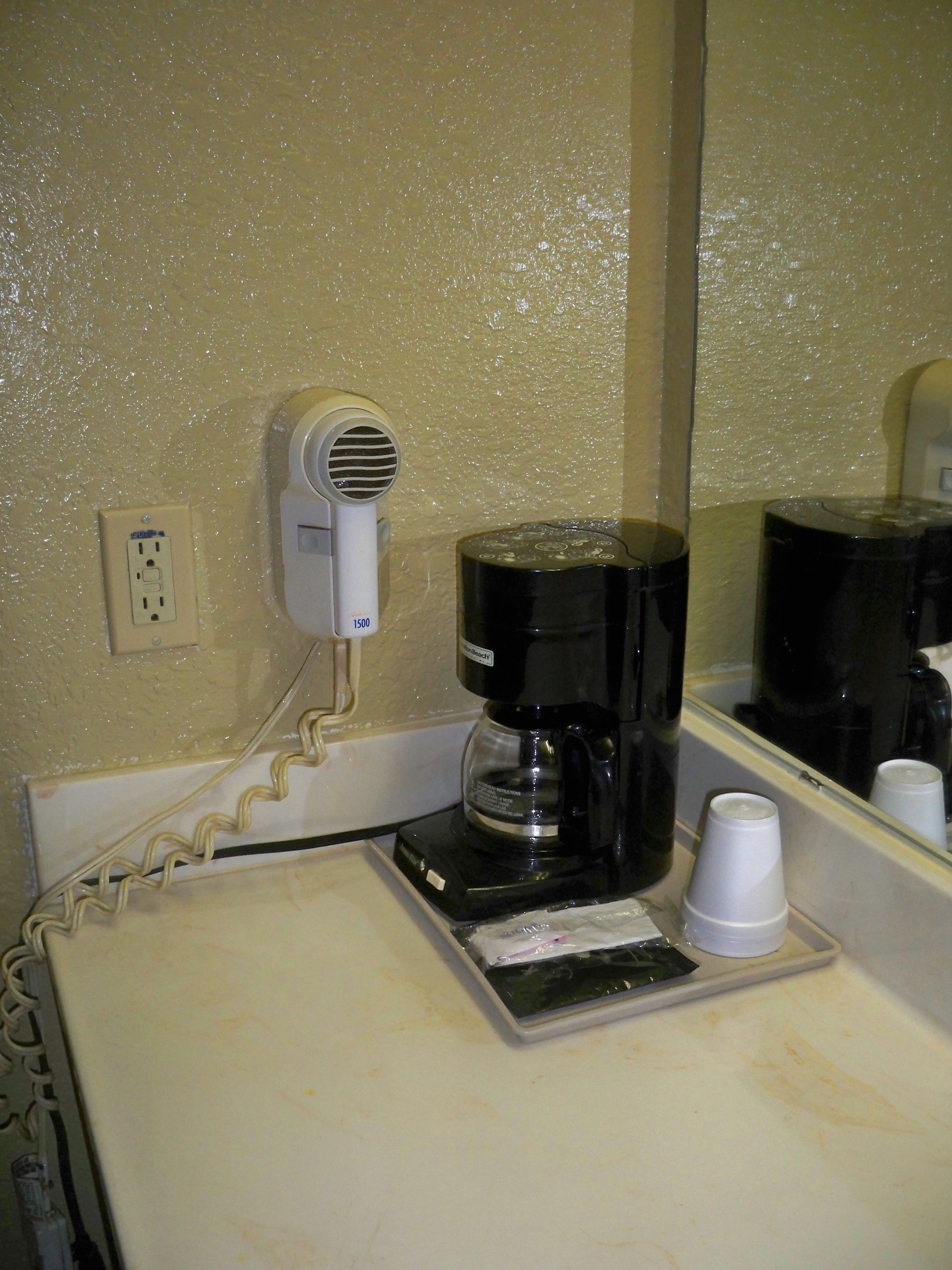 A hotel room with a coffee maker on the counter, a hair dryer on the wall, and a mirror reflecting the coffee maker and a cup.