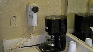 A hotel room with a coffee maker on the counter, a hair dryer on the wall, and a mirror reflecting the coffee maker and a cup.
