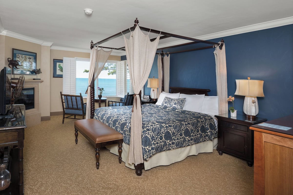 A four-poster canopy bed with a patterned bedspread, a wooden bench, a nightstand with a lamp, and a view of the ocean through a window.