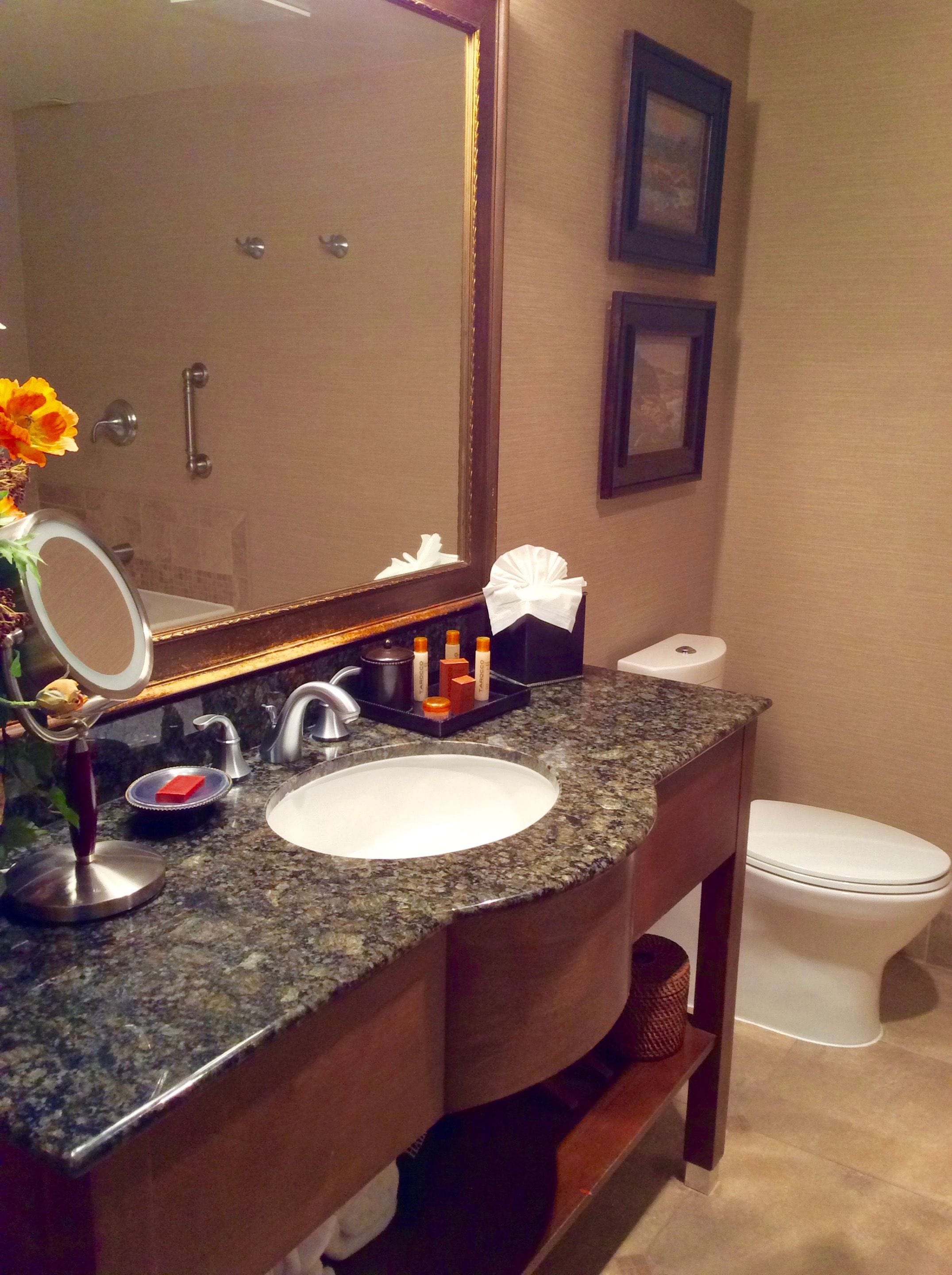 A bathroom with a granite countertop, a white sink, a mirror, a towel dispenser, and a toilet.