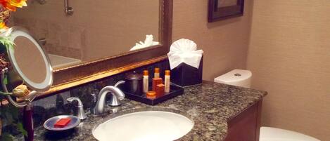 A bathroom with a granite countertop, a white sink, a mirror, a towel dispenser, and a toilet.
